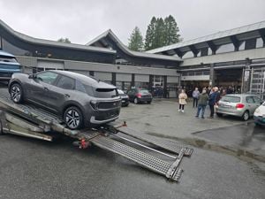 Den nye bilen vår (den grå) på veg nedover rampa på transportbilen utanfor bilverkstadhallen på skulen. Ei gruppe elevar og vaksne følger med i bakgrunnen